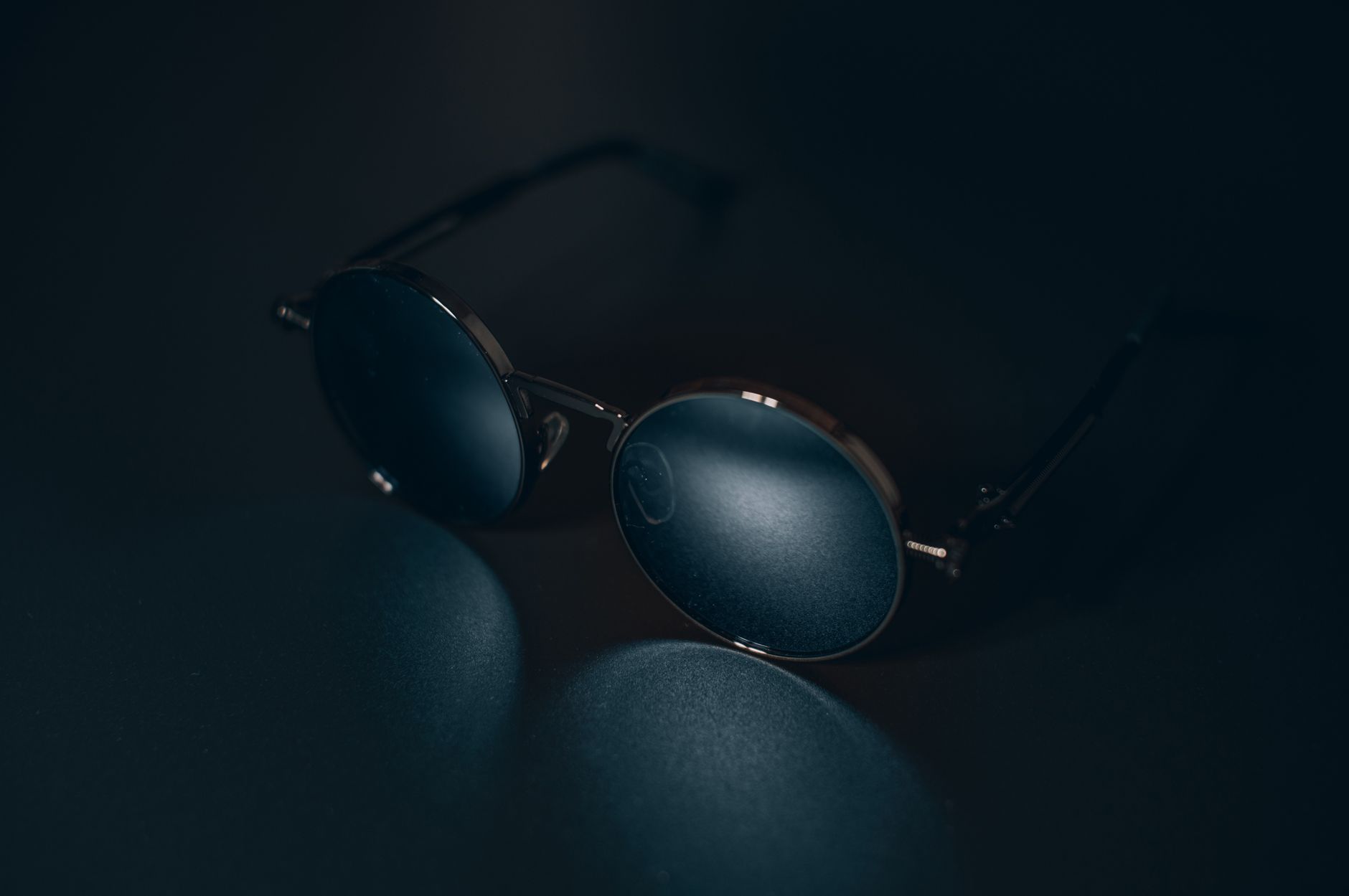A pair of round metal-framed sunglasses resting on a dark surface, lit from one side so the rims and the dark lenses catch the light.