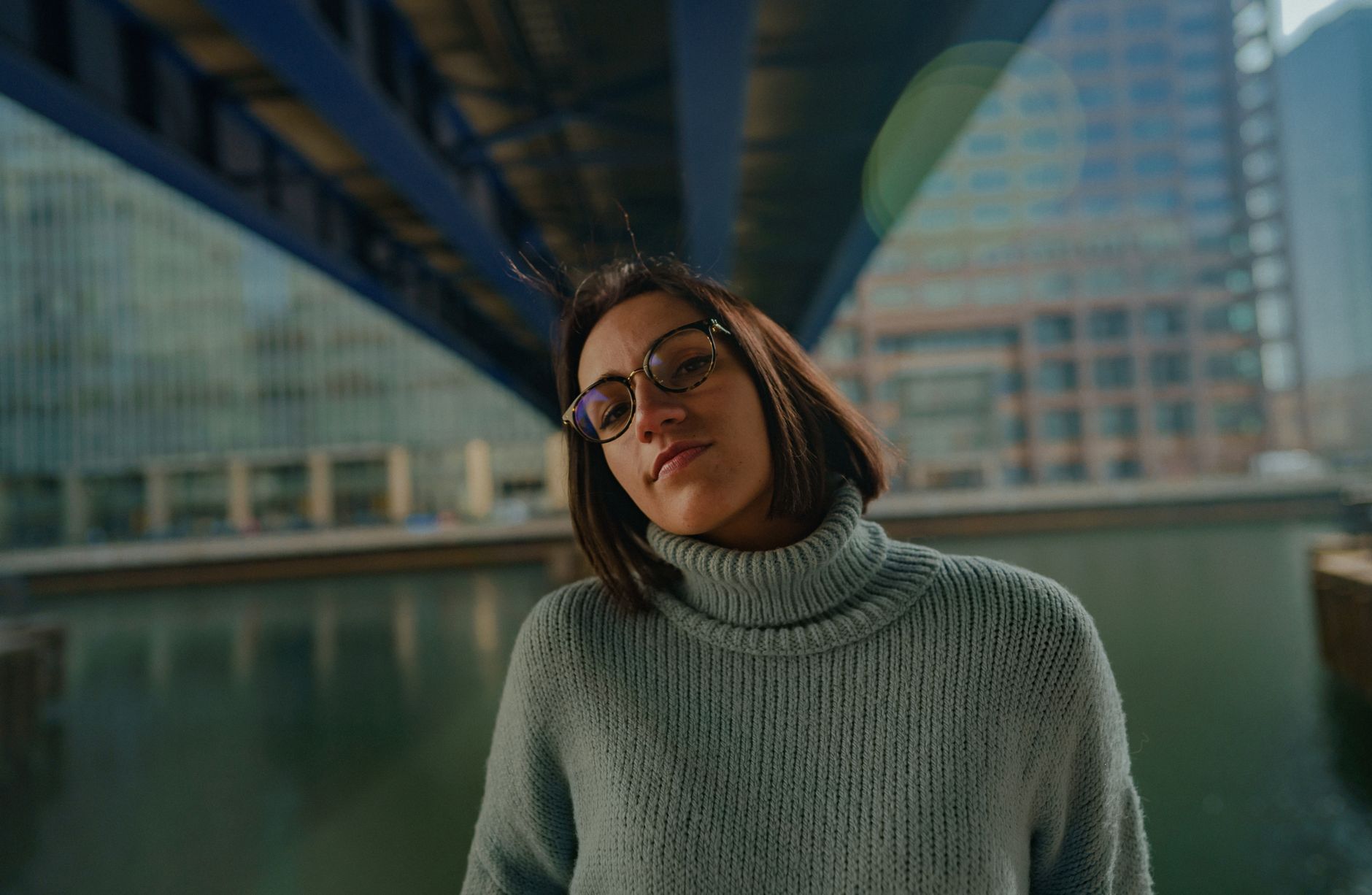 Portrait of a woman wearing a turtleneck sweater and glasses, standing under a bridge by the water. — illustrating What Is a Saddle Bridge, and What Problem Does It Solve?