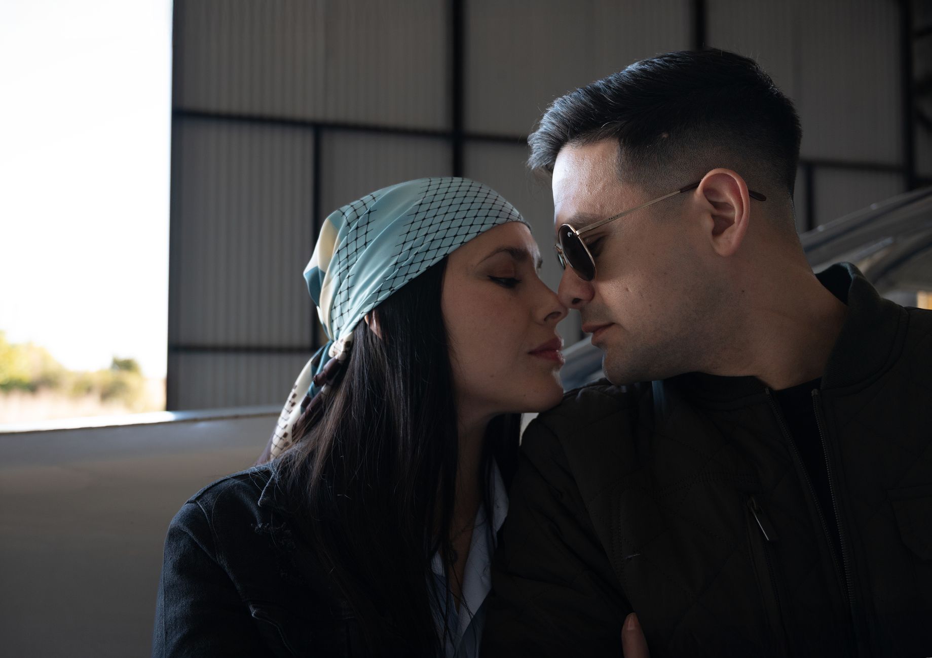 Romantic pose of couple in sunglasses and scarf inside a hangar. — illustrating What Is the Story Behind the Aviator Sunglasses Shape?