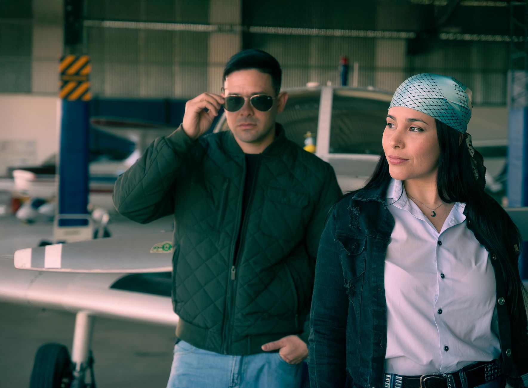 Two individuals in a hangar, sunglasses and casual attire, near aircraft. — illustrating What Is the Story Behind the Aviator Sunglasses Shape?